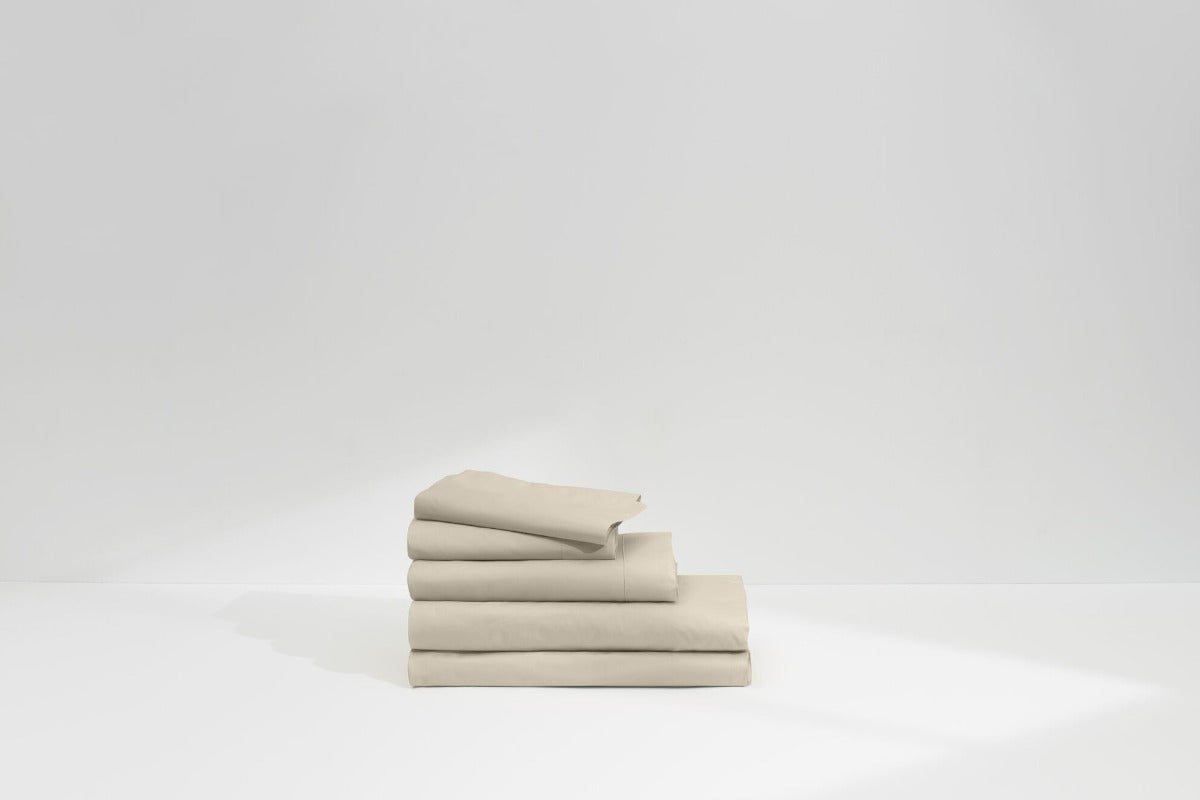 Stack of neatly folded beige bed sheets on a plain white surface with a minimal background.