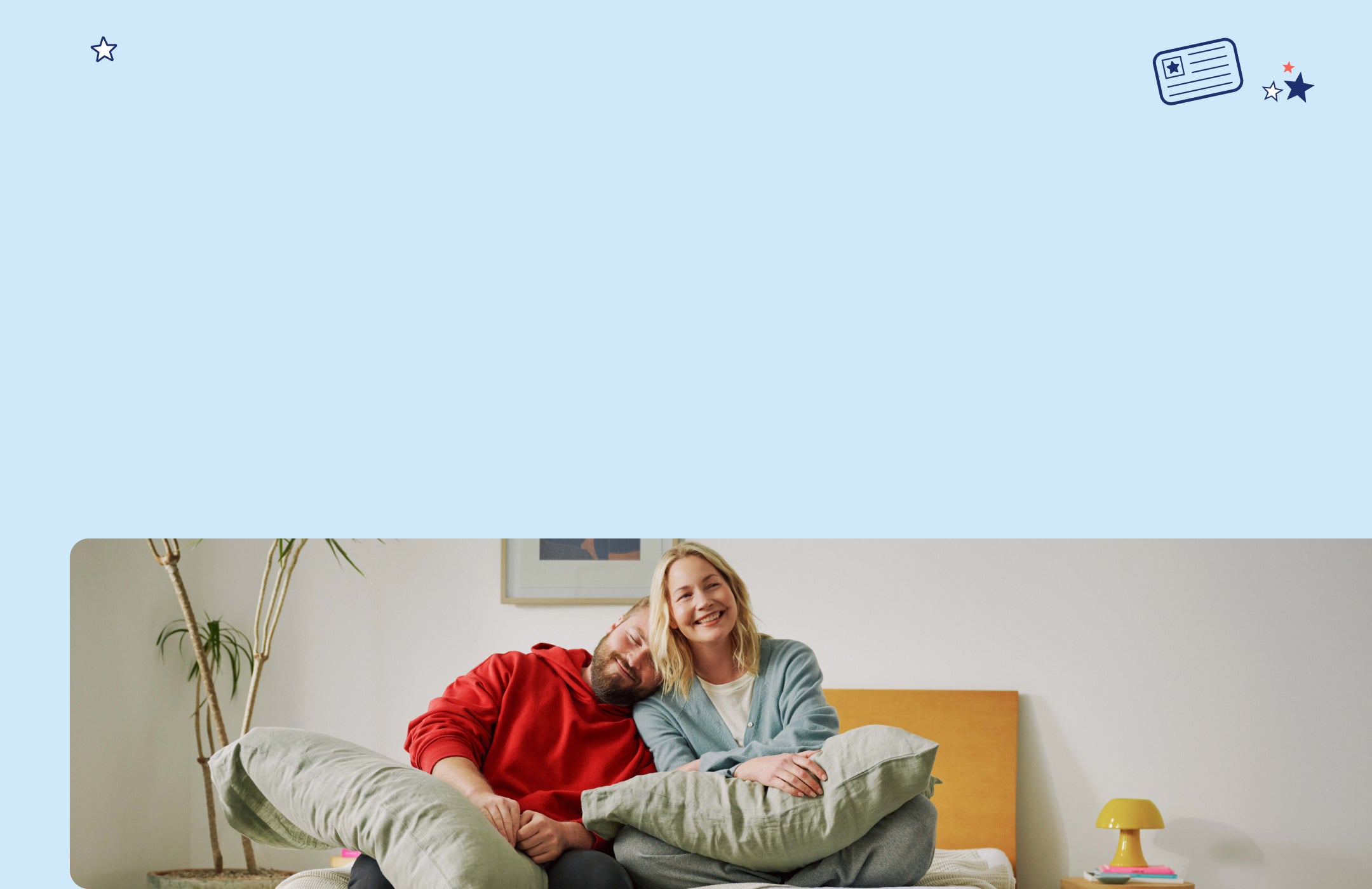 A couple sits on their bed, smiling, with pillows on their laps.