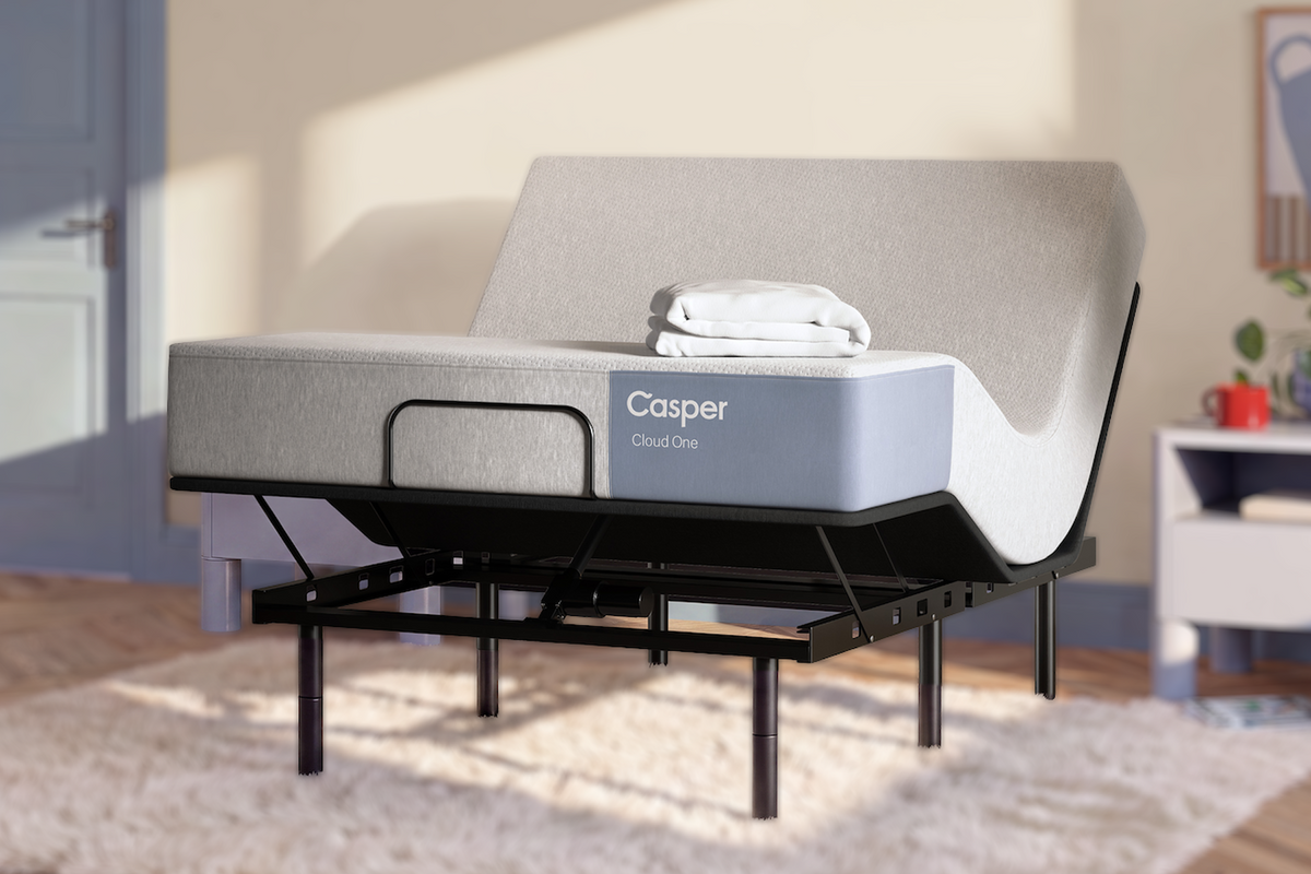 A Casper Cloud One mattress on an adjustable bed frame with a light gray upholstered mattress and a black metal support base, shown in a styled bedroom setting with warm sunlight and a white rug.