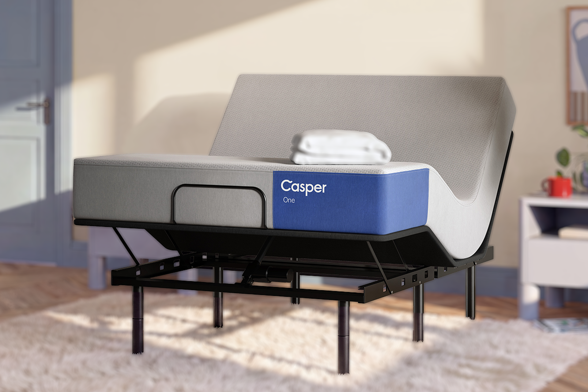 A Casper One mattress on an adjustable bed frame with a gray mattress and white folded bedding, shown in a styled bedroom setting with warm sunlight and a rug.