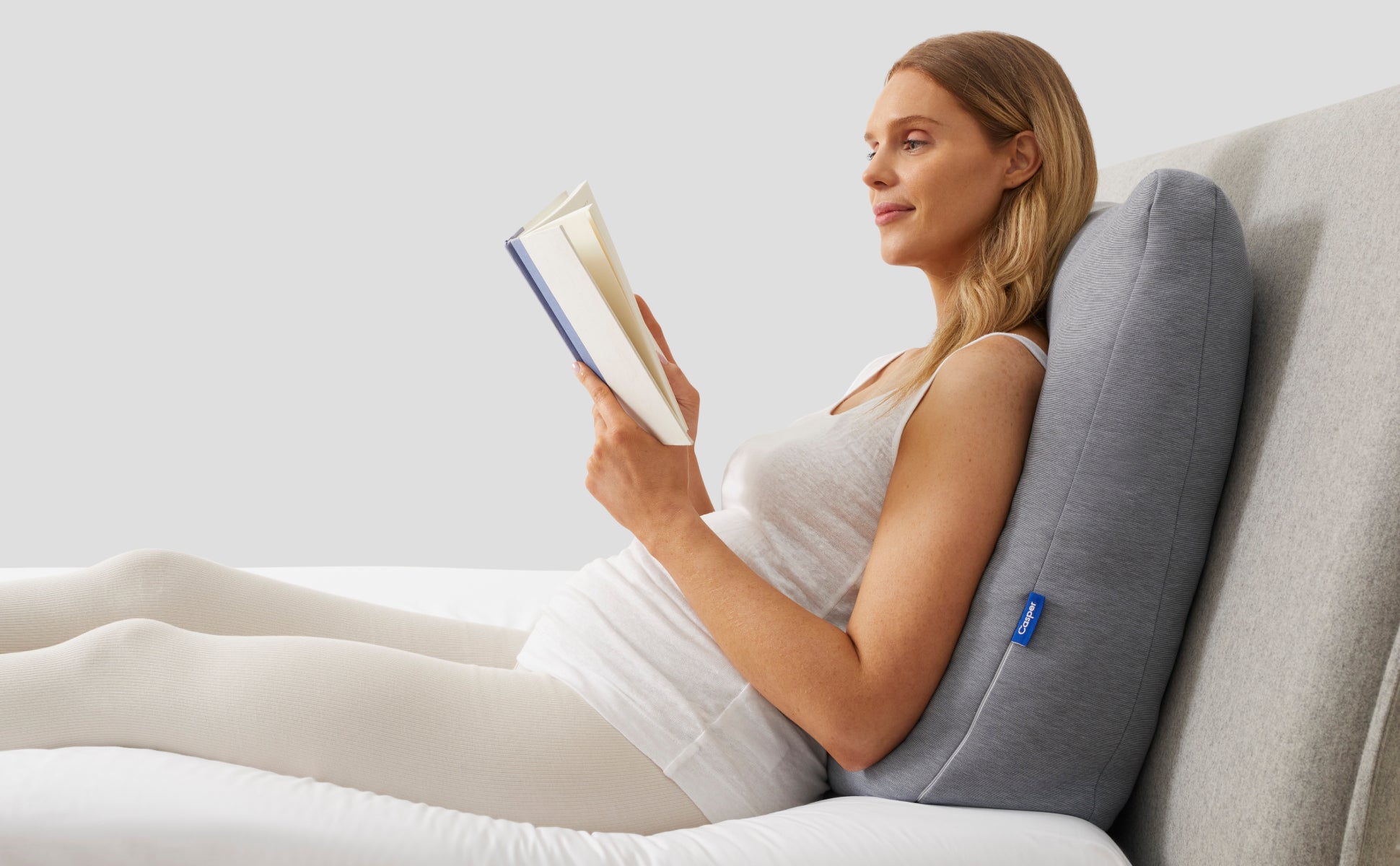 A person leaning back on the Casper Backrest Pillow and reading in bed.
