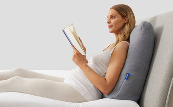 A person leaning back on the Casper Backrest Pillow and reading in bed.
