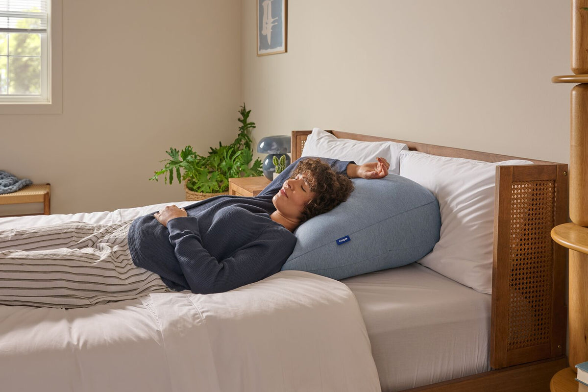 A woman leans back against the Backrest Pillow on a bed.