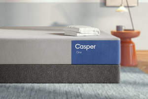A Casper One mattress shown in a styled bedroom setting, featuring a white and blue top section with the brand name 'Casper One' printed on the side, and a dark gray upholstered base. A folded white pillowcase rests on top.