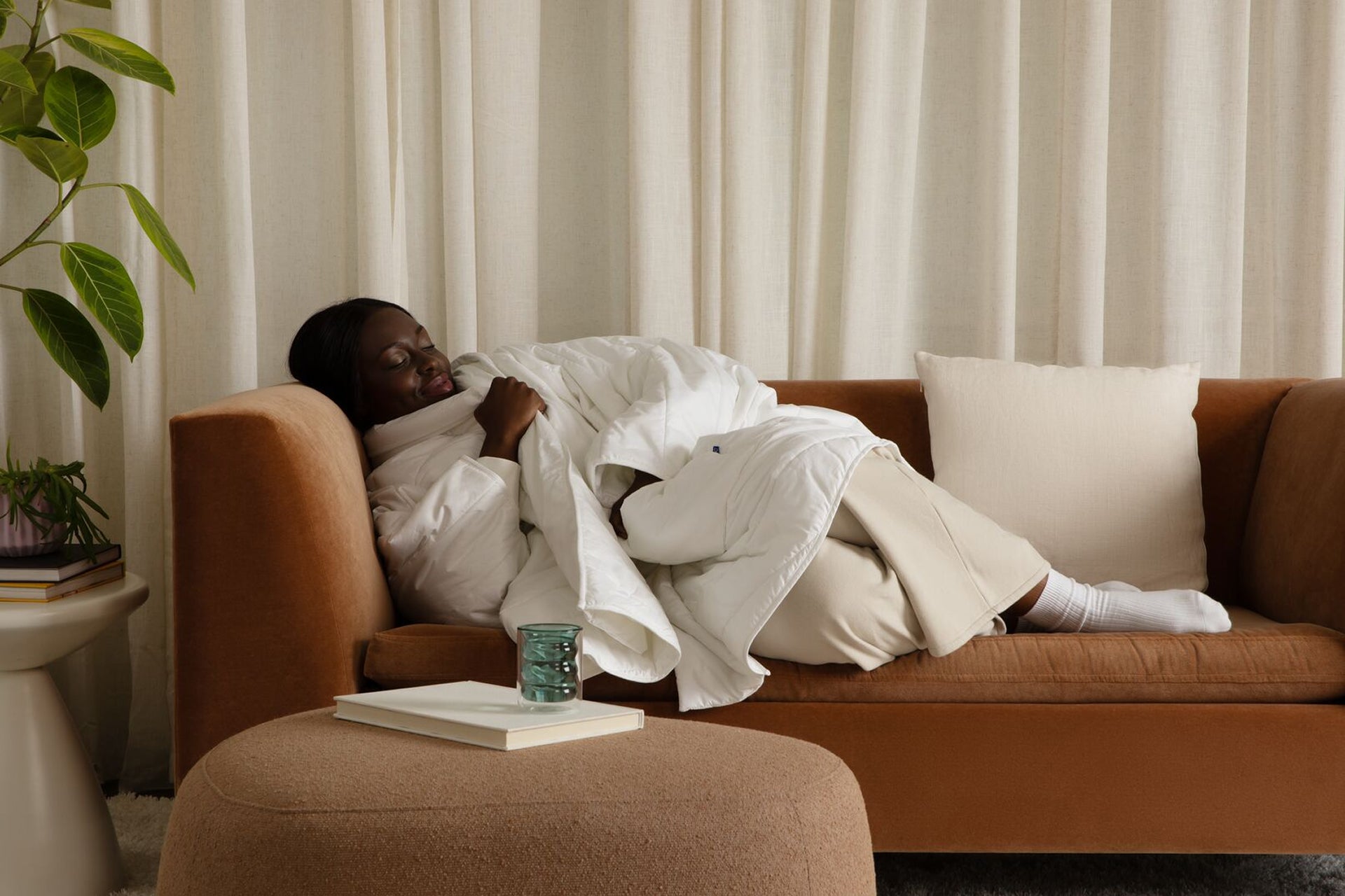A person reclines on a brown sofa, wrapped in a white Blanket Robe, in a softly lit living room with cream curtains, a plant, and a small ottoman in the foreground.