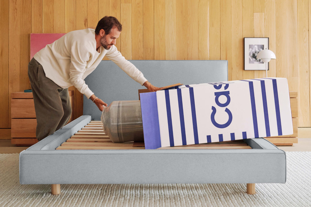 A man unboxes his new Casper mattress on top of an empty bed frame.