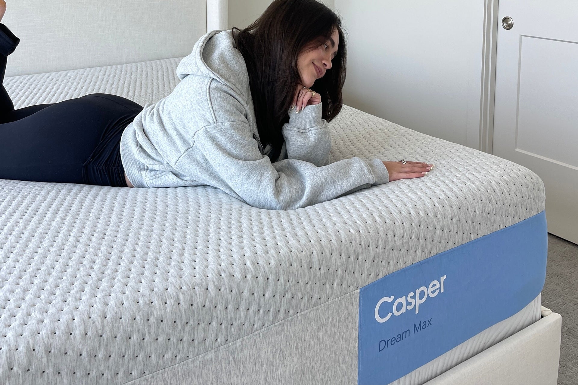 A woman in a light gray hoodie and black pants lies on her stomach on a white Casper Dream Max mattress in a bedroom setting.