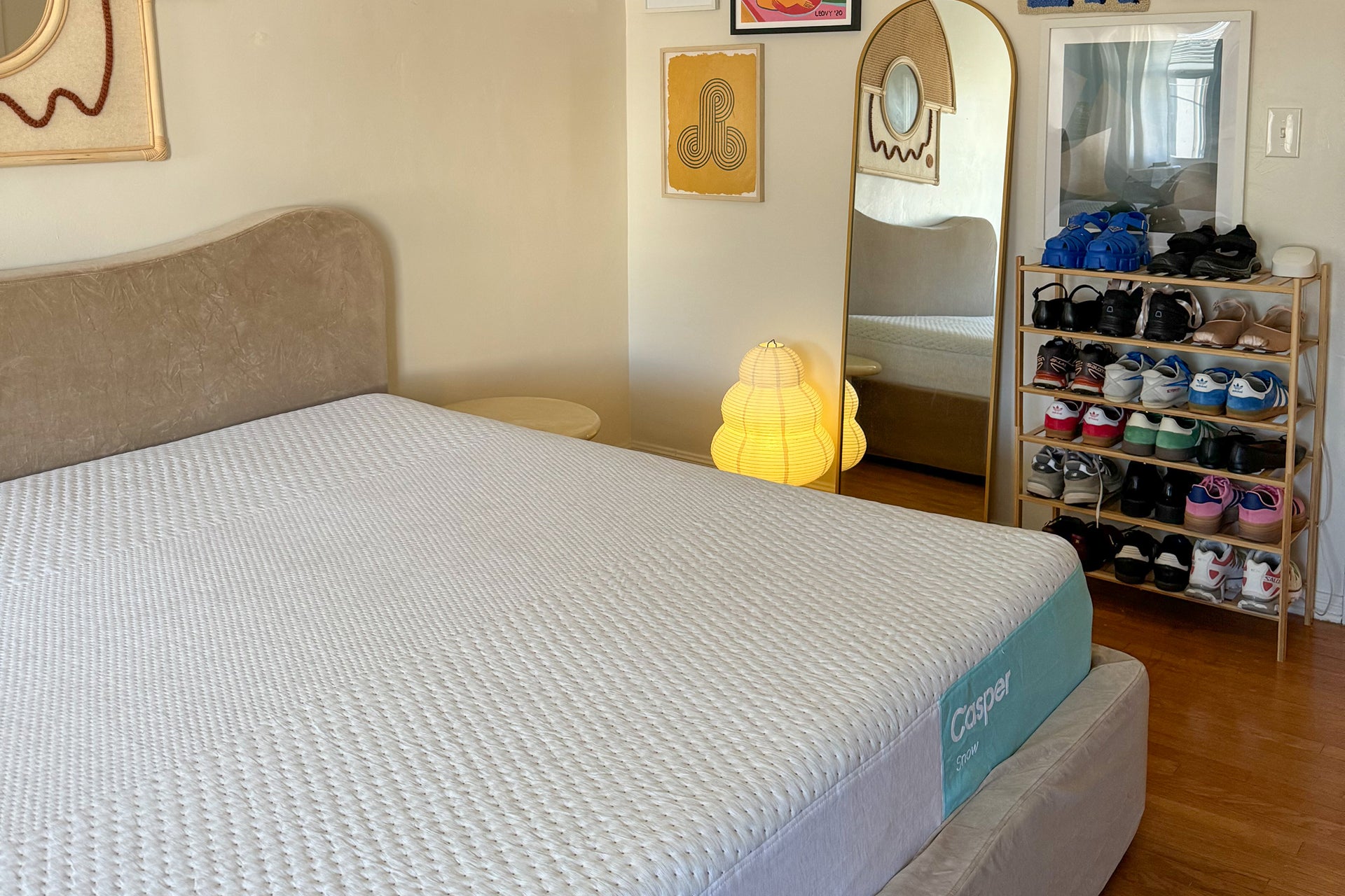 A bed with a Casper Snow mattress in a bedroom setting, featuring a beige upholstered headboard, a yellow glowing lamp, a tall mirror, and a wooden shoe rack filled with shoes