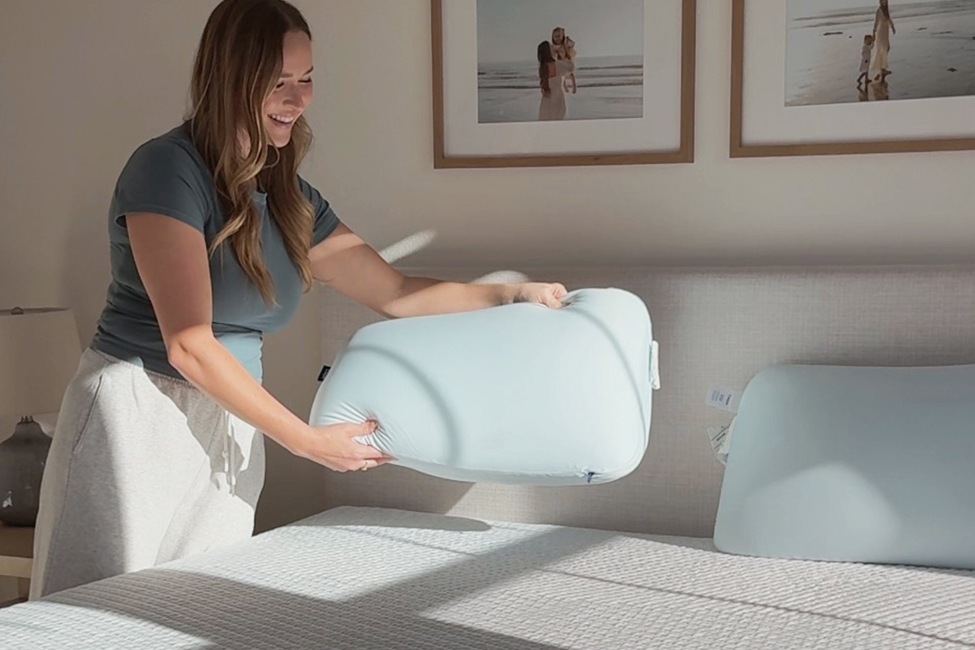 A woman in a bedroom holds a light blue contoured pillow, with another matching pillow resting on the bed beside her.