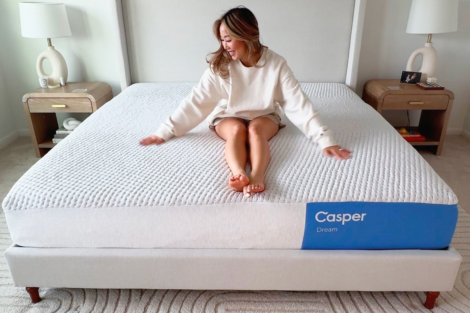 A woman sits on a white Casper Dream mattress in a bed frame, wearing a cream cardigan and shorts in a bright bedroom setting with bedside tables and lamps.
