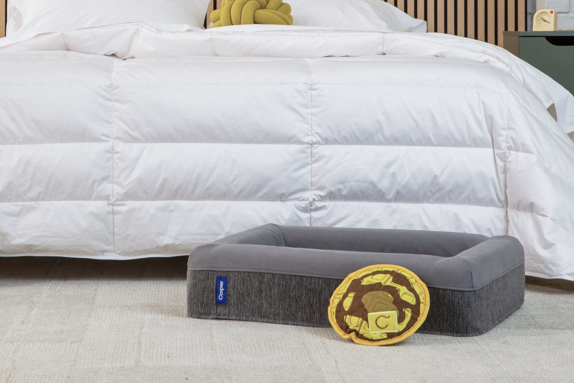 A gray rectangular pet bed with a gold foam toy, positioned on a light carpet in front of a bed with white bedding