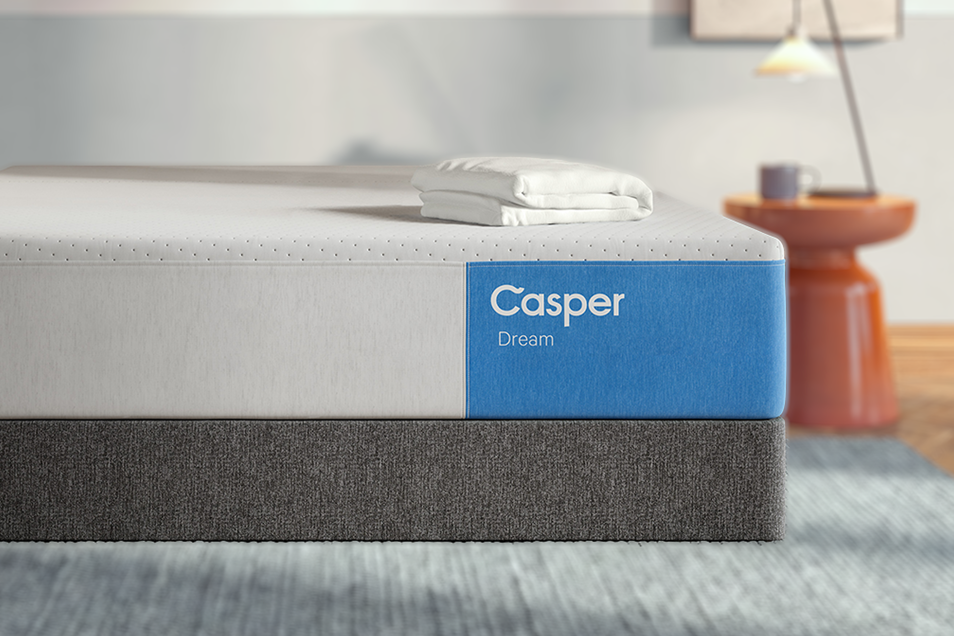 A Casper Dream mattress shown in a styled bedroom setting, featuring a white top section, a blue side label with the white Casper brand name, and a dark gray upholstered base.