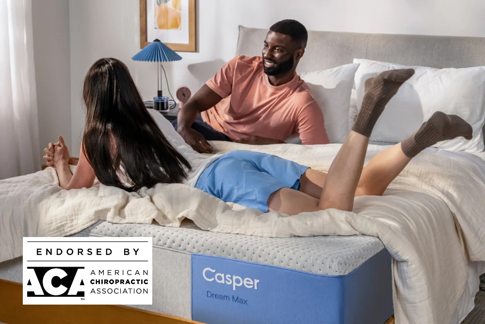 A couple lies happily in bed during the daytime. One is leaning on the edge of the bed, and the other is lying on their stomach but pressed up on their elbows.