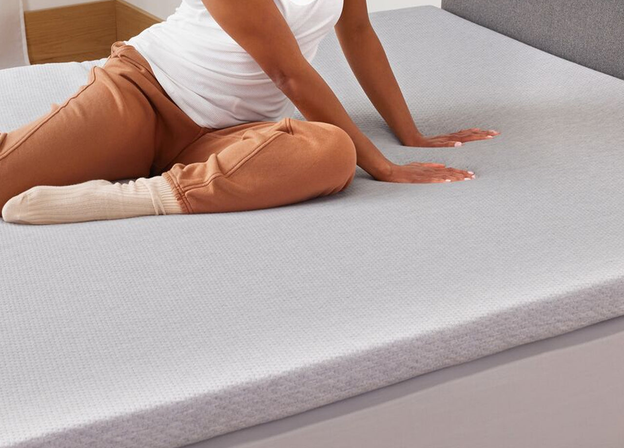 A peson sits on their Essential Topper in bed, feeling the comfort of the mattress topper with their hands.