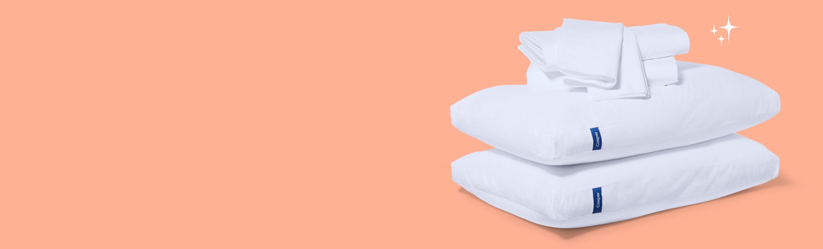 A stack of two pillows, folded sheet set, and pillowcases against a faded salmon color.