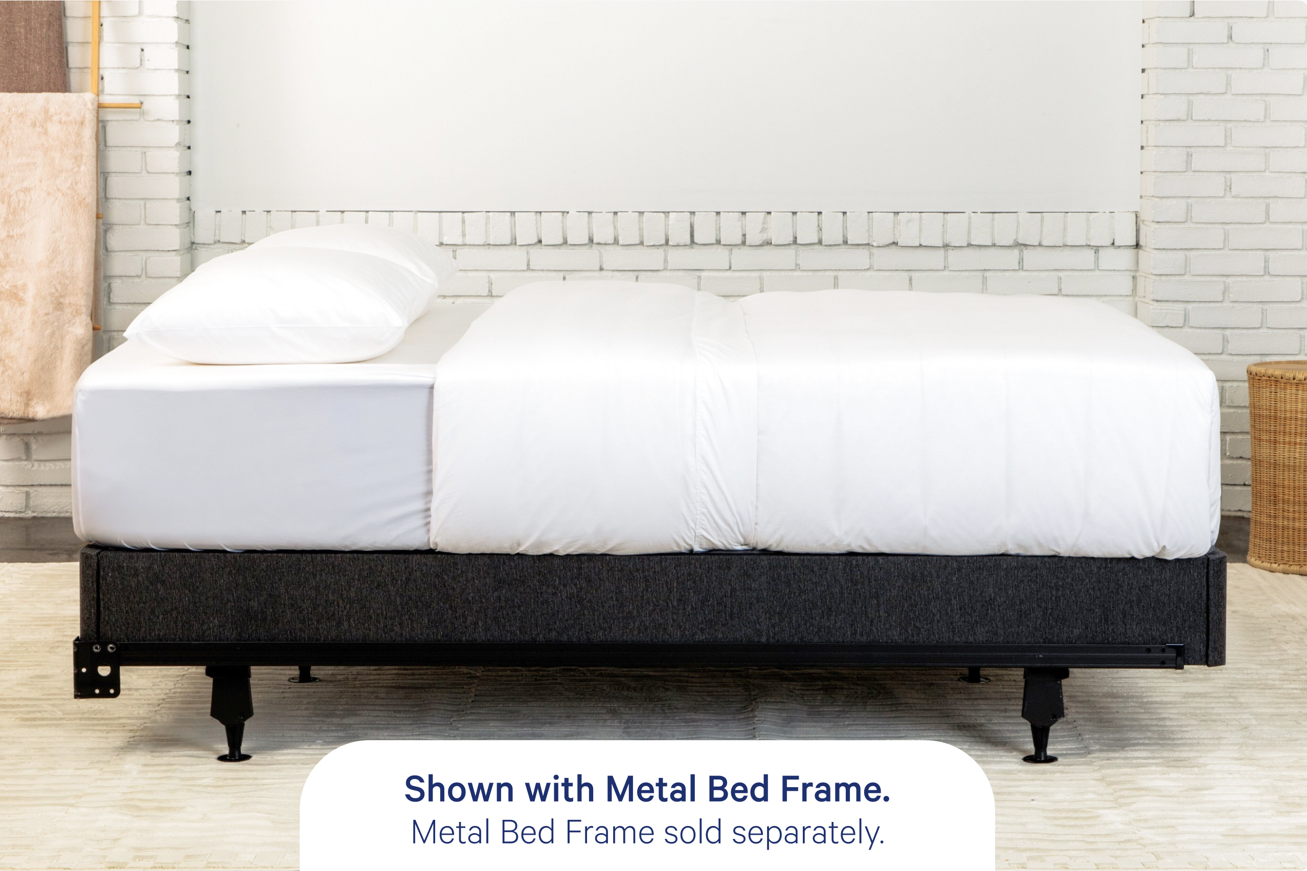 A bed with white bedding and pillows shown on a box spring, Casper's Foundation, placed on a dark metal bed frame in a bright room with a white brick wall. Text at the bottom reads, 'Shown with Metal Bed Frame. Metal Bed Frame sold separately."