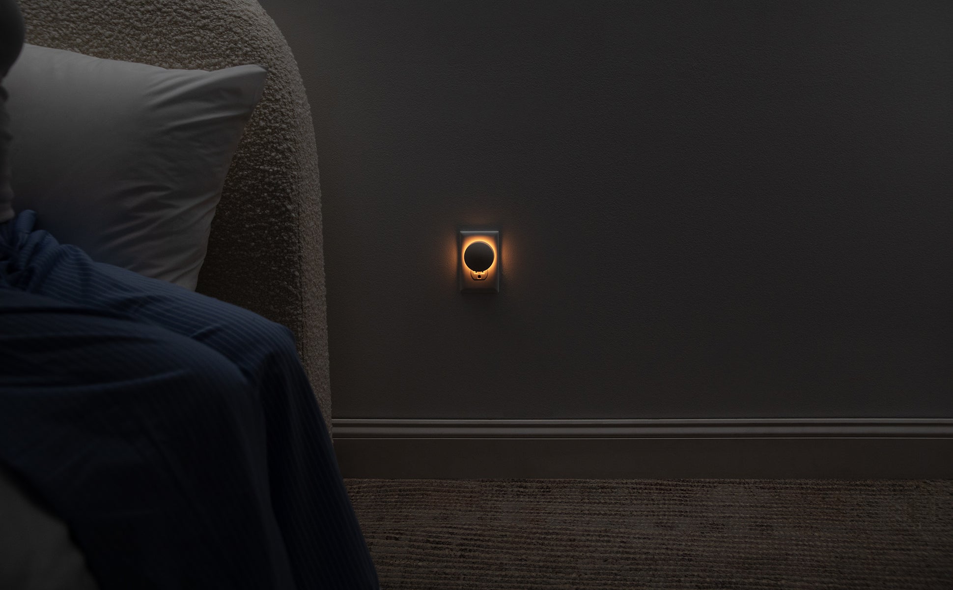 Image of the Glow Nightlight, plugged into a wall, on Sleep Mode next to a bed