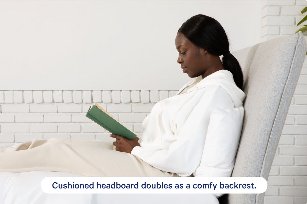 Haven Bed Frame, gray, side view of a woman lying back against the headboard with a book. Cushioned headboard doubles as a comfy backrest.