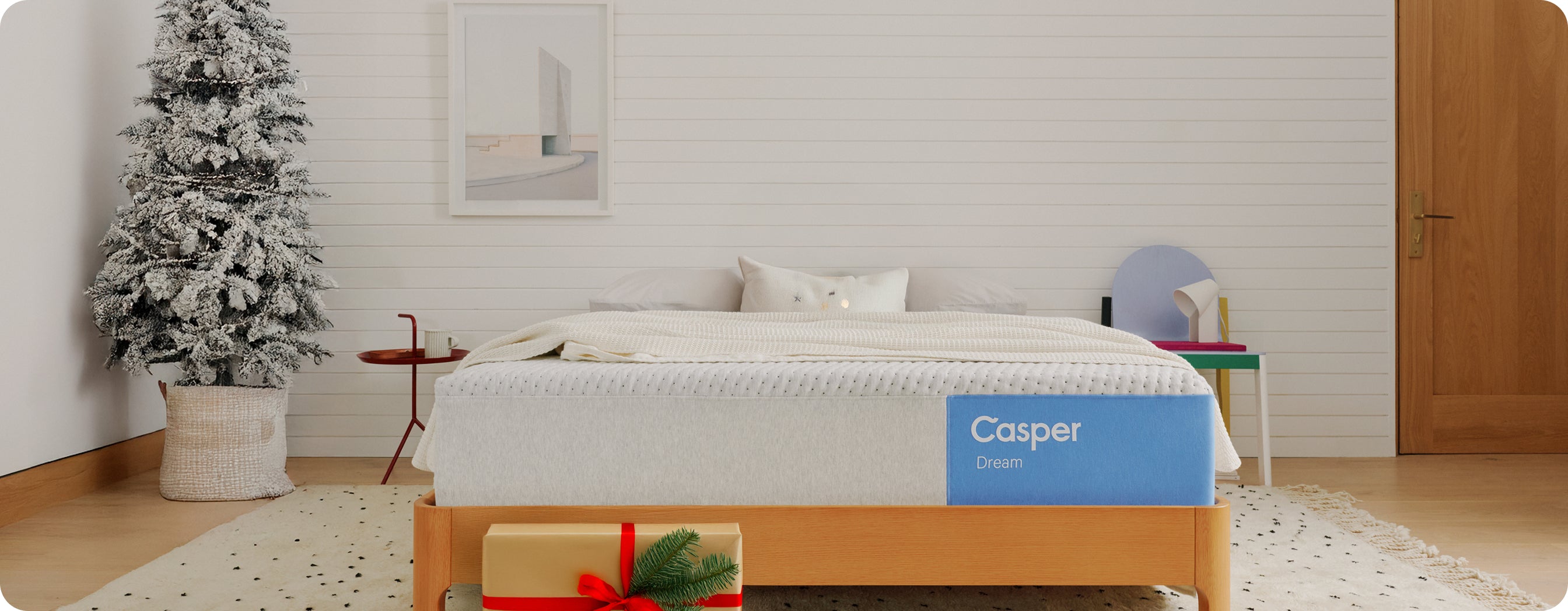 A Casper Dream mattress is on a wooden bed frame in a bedroom with a Christmas tree in the corner and a wrapped gift on the floor in front of the bed.