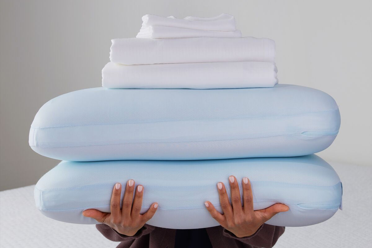 A person holding a stack of two pillows and a folded sheet set