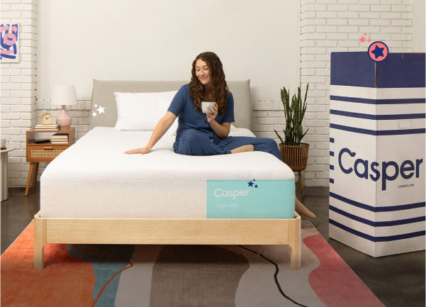 A woman sitting in bed on a Casper Snow Max mattress. The mattress delivery box with the word Casper is upright on the floor next to the bed.