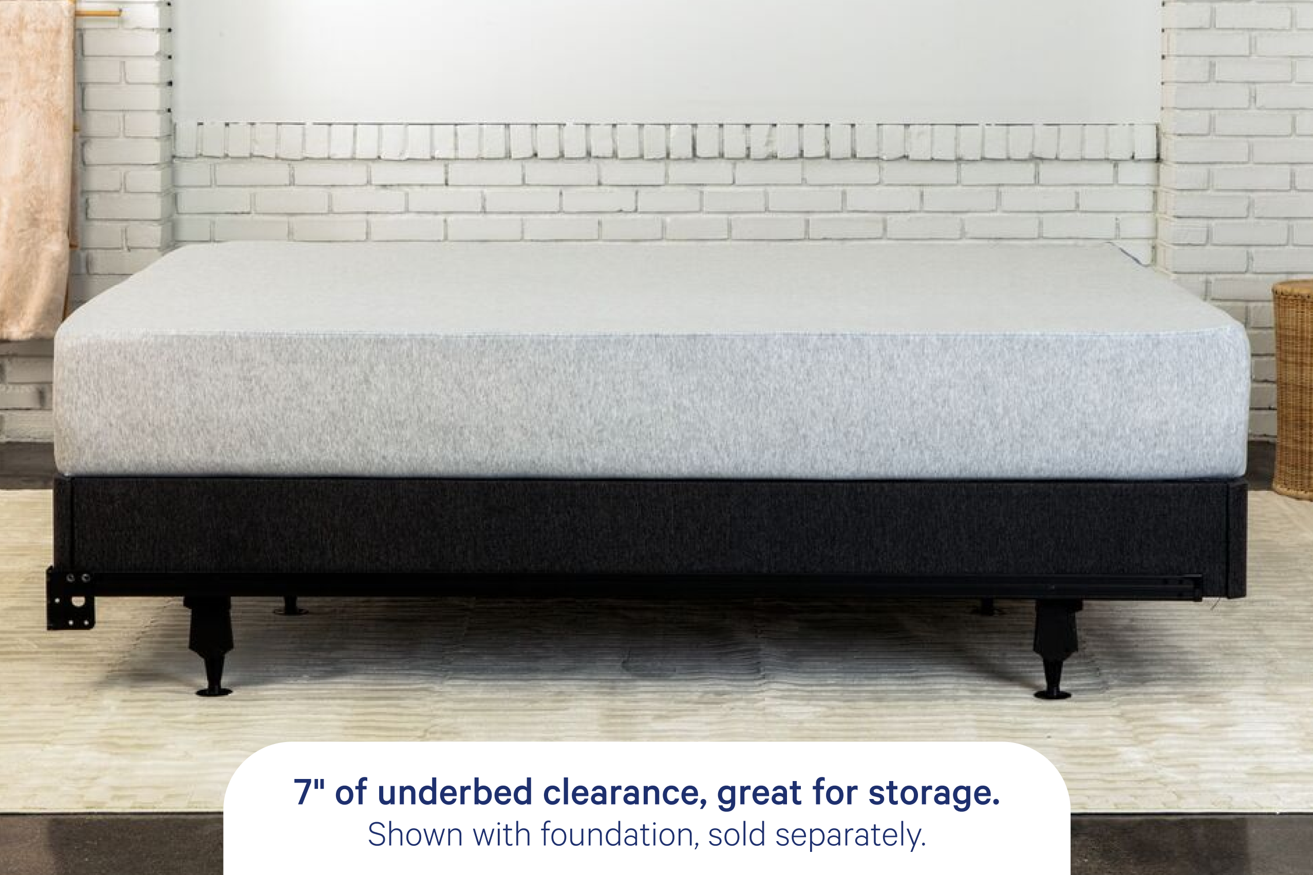 A bed frame with a light gray mattress shown in a styled bedroom setting with a white brick wall and rug. A text overlay at the bottom reads, '7" of underbed clearance, great for storage. Shown with foundation, sold separately.'
