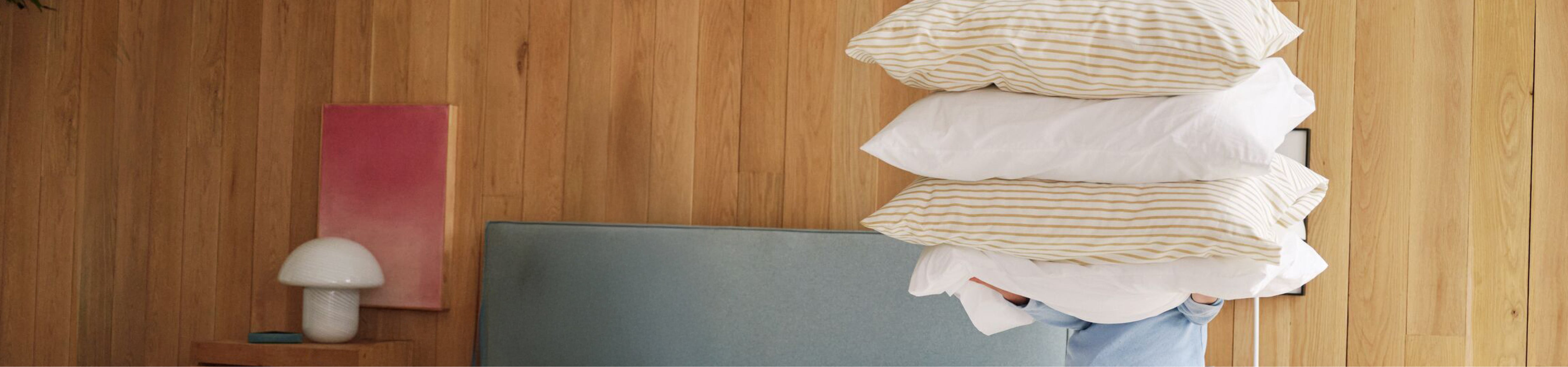 A person standing in a bedroom holds a stack of four pillows.