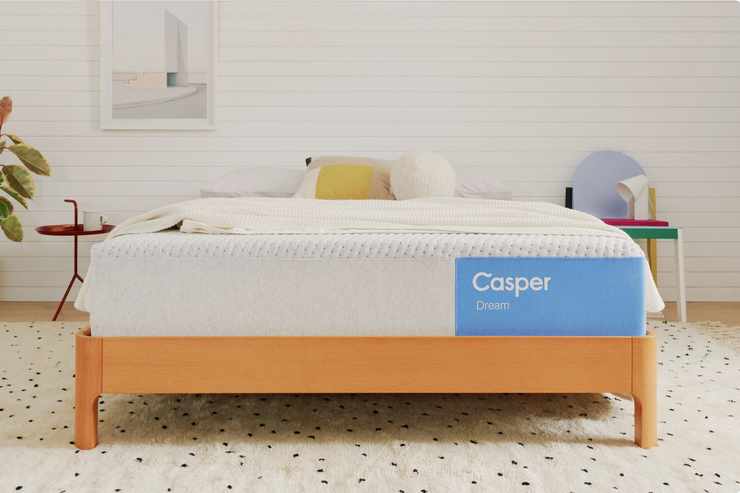 A bed with a white Casper Dream mattress on a wooden bed frame, styled in a bright, modern bedroom with a white shiplap wall, colorful side tables, and a patterned rug.