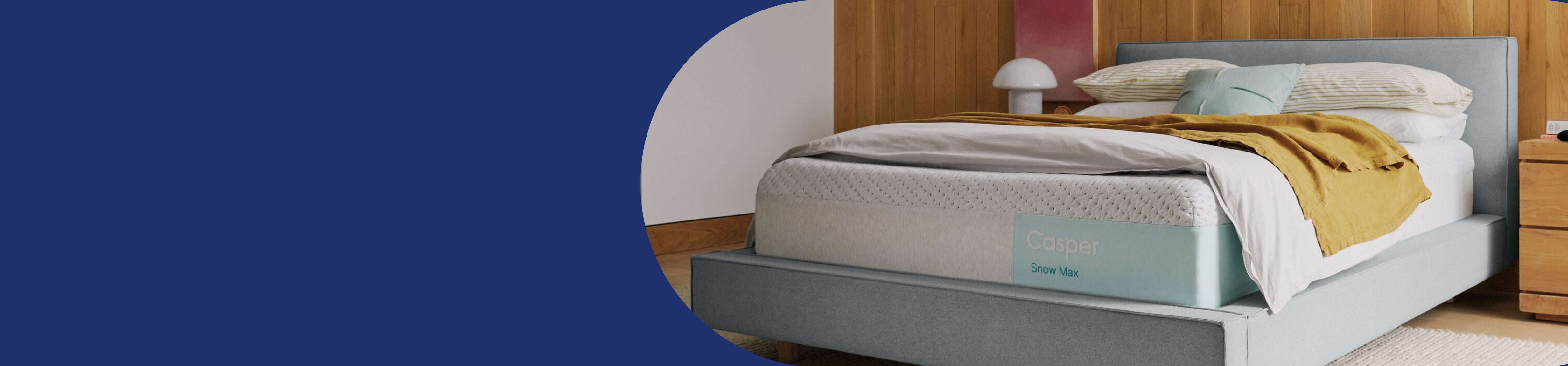 Casper' Snow Max mattress on a fabric bed frame with a headboard. Two blankets and multiple pillows are on top of the bed.
