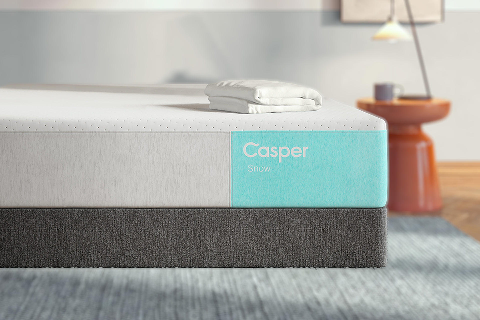 A Casper Snow mattress shown in a styled bedroom setting, featuring a white and light gray top section with a turquoise side label and a dark gray textured base.