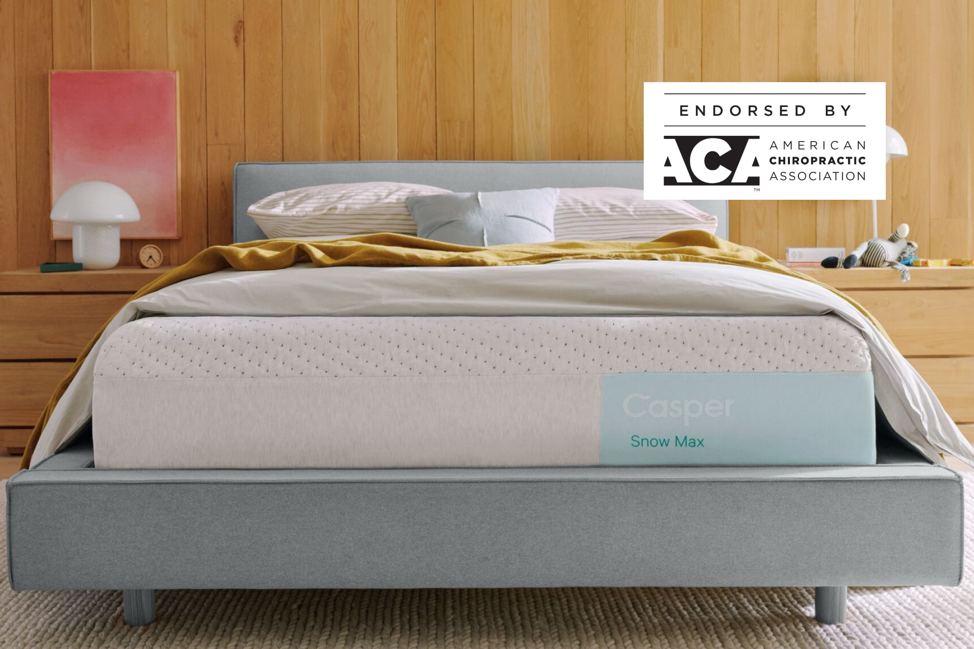 Casper's Snow Max mattress on a bed frame with pillows and a blanket folded on top. A badge on the image indicates that this mattress is endorsed by the American Chiropractic Association.