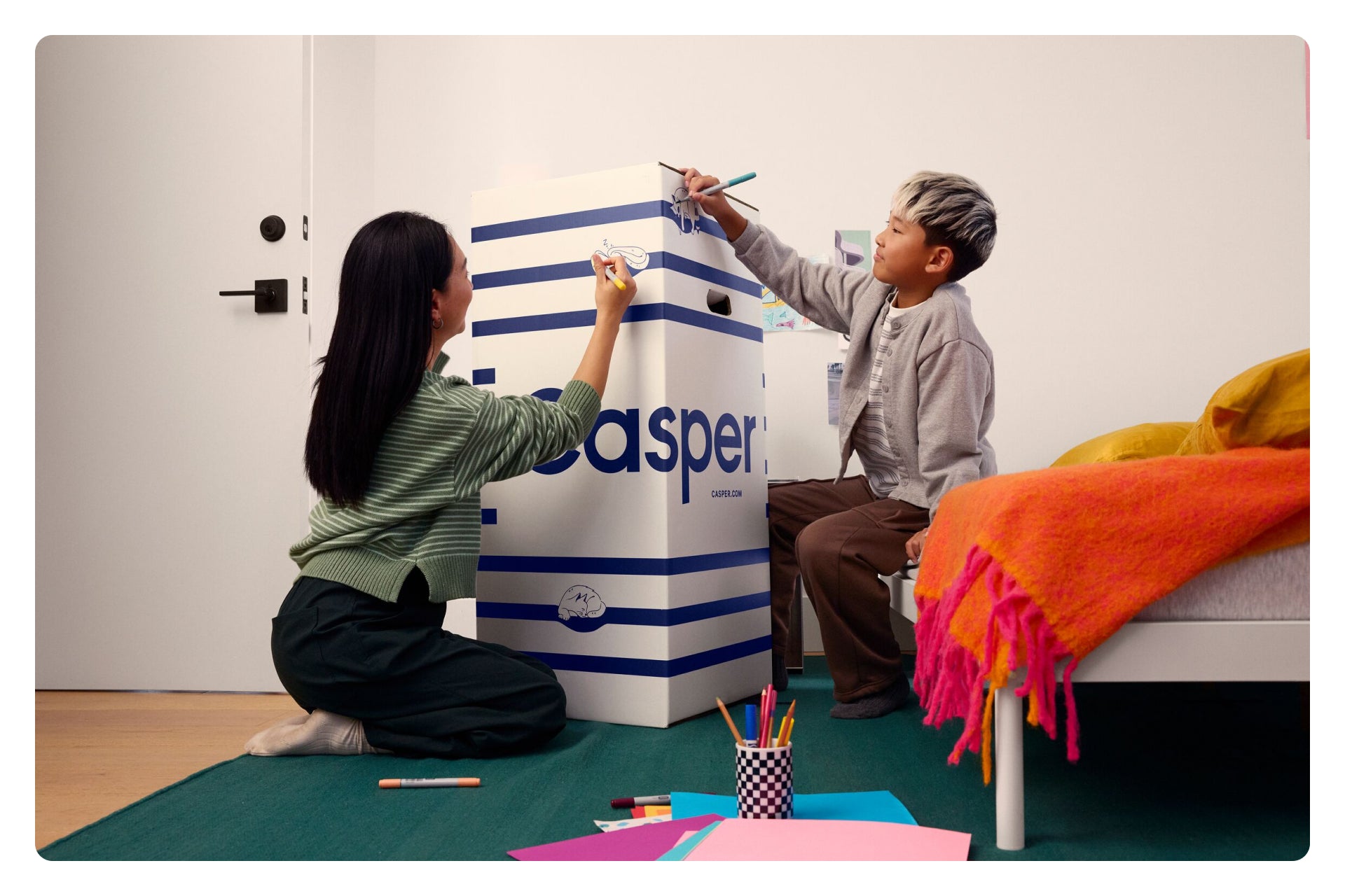 Two people painting a large 'Casper' branded box in a room with furniture and decor.