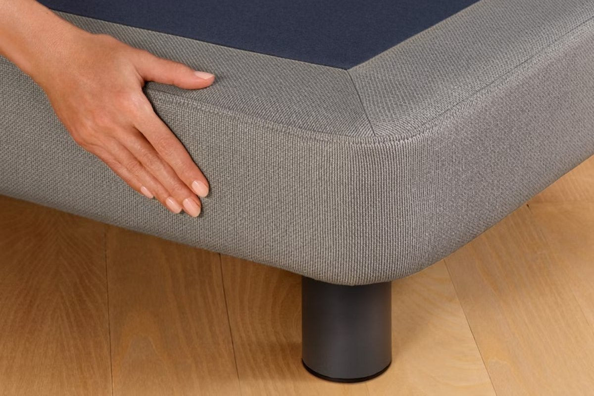 A person's hand feels the fabric near the corner of the Upholstered Bed Frame, gray color.