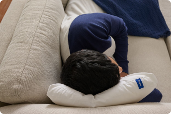 A man takes a nap on a couch, lying on his side on a Nap Pillow.