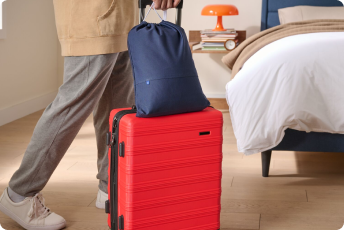 A person walks their small, rolling suitcase with a Nap Pillow in its travel bag on top of it.
