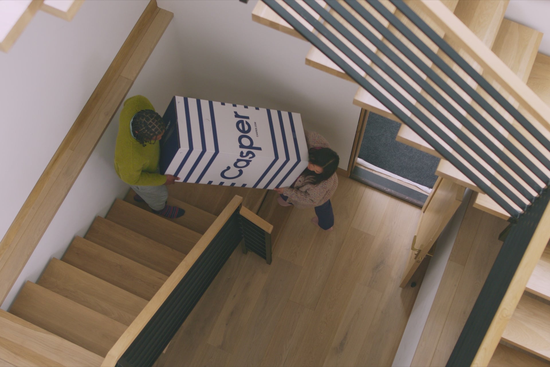 A couple carries their new Casper Dream mattress upstairs in its delivery box. Once in the room, a woman is seen lying comfortably on the new mattress.