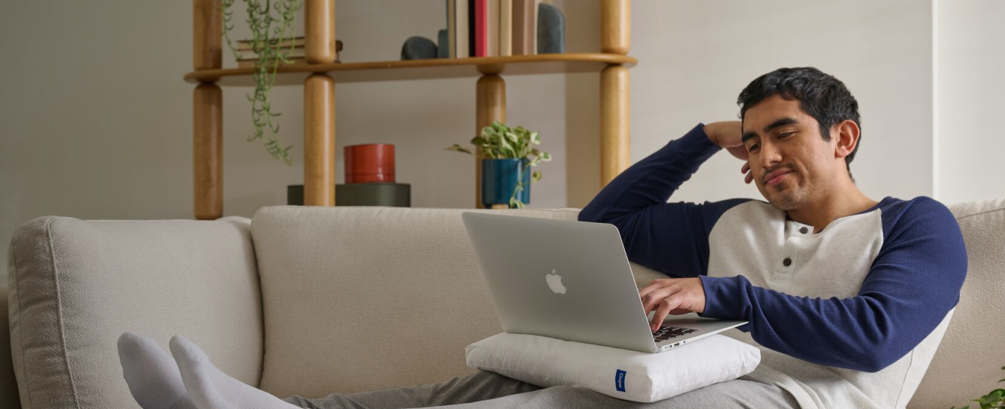 Man using a laptop on a couch in a cozy living room. The laptop sits atop his Mini Nap Pillow on his lap.