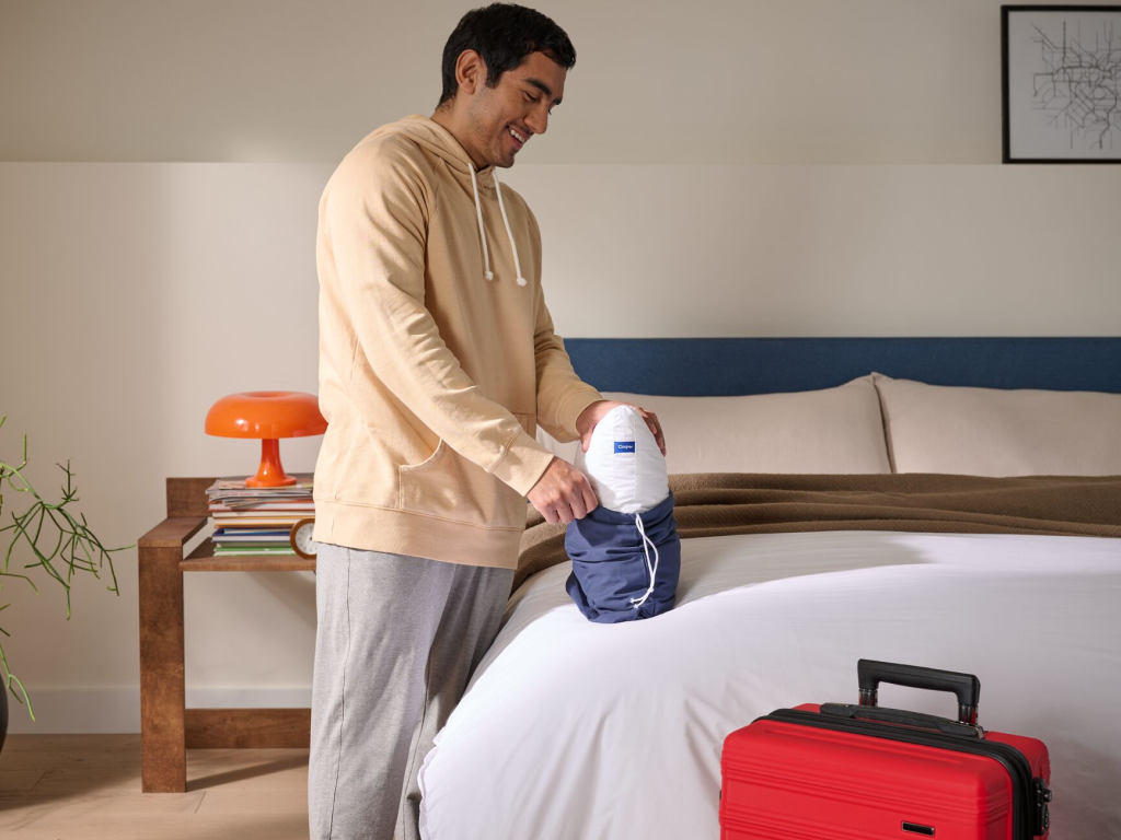 Man standing besides a bed putting his Mini Nap Pillow into its small carrying bag for travel.
